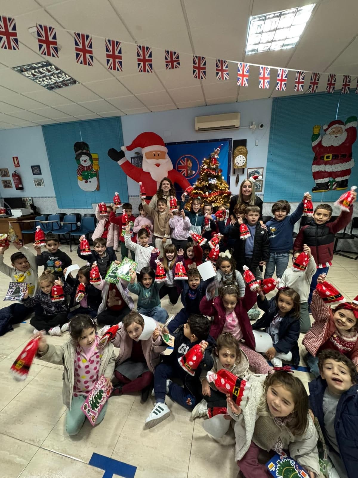 Los alumnos de Primaria levantan sus gorros de Papá Noel con sus profesoras en la fiesta de Christmas